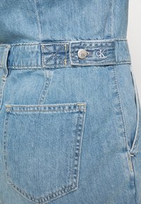 Světle modré džíny z denimu s viditelným šitím, logem CK na pásku a zadní kapsou. Obsahují zapínání na knoflík a zip.