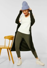 Długi czarny kardigan, biały t-shirt, oliwkowe legginsy i kremowe sneakersy na wysokim obcasie. Modelka nosi jasnoniebieską wełnianą czapkę. Obok drewniane krzesło.
