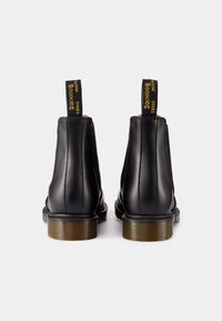 Černé kožené kotníkové boty s elastickými bočními díly. Mají žlutou poutko a texturovanou hnědou gumovou podrážku. Podél okrajů jsou viditelné švy.