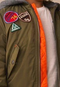 Veste vert olive avec capuche grise et doublure orange, ornée de plusieurs écussons brodés en bleu, marron et vert. Fermeture éclair et poche à pression.