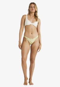Bílý bikiny top z texturované látky s širokými ramínky, spárovaný se zeleným vzorovaným bikinovými spodky s vysokým střihovým výřezem. Jednoduchý a funkční design.