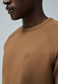 Sudadera marrón con cuello redondo, puños acanalados y una etiqueta cuadrada que dice "SALSA JEANS" cosida en el pecho. Textura de tela suave.