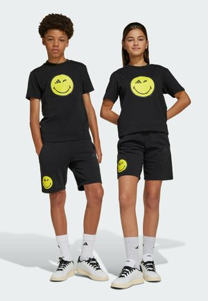 T-shirts en coton noir et shorts avec un imprimé de smiley jaune vif. Baskets avec des lacets noirs, accompagnées de chaussettes rayées blanches.