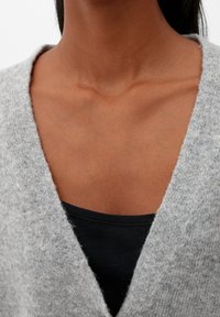 V-ringad tröja i ljusgrått stickat material med en mjuk textur. Under finns en svart linne som syns och kompletterar tröjans design.