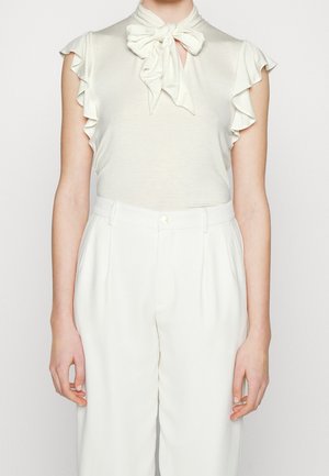 Blouse pâle sans manches avec emmanchures volantées et nœud noué au cou, associée à un pantalon blanc plissé taille haute.