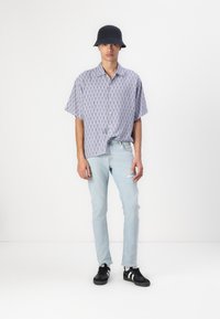 Camisa de manga corta con estampado en azul claro y blanco, combinada con jeans rasgados azul claro y sombrero tipo cubo negro, llevada con zapatillas negras.