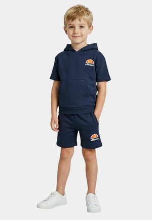 Jeune garçon debout, portant un sweat à capuche sans manches bleu marine et un short assorti avec des logos ellesse orange et blancs, baskets blanches.