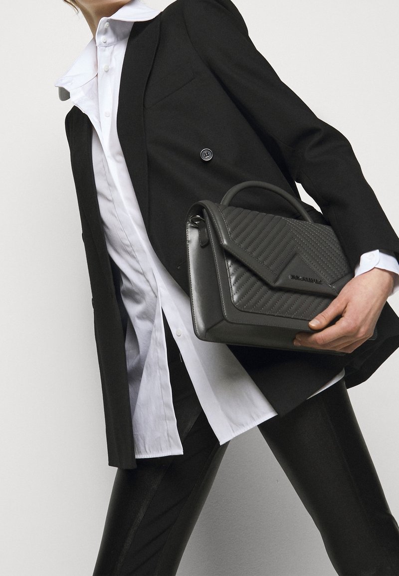 Blazer noir porté sur une chemise blanche à col, tenant un sac à main en cuir gris avec un motif matelassé et une poignée supérieure. Pantalon noir slim visible.
