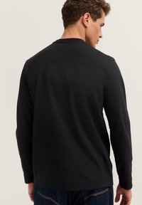 Mann trägt ein einfaches schwarzes Langarmhemd und dunkelblaue Jeans, steht mit dem Rücken zur Kamera vor einem neutralen Hintergrund.