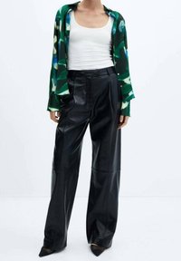 Donna che indossa pantaloni in pelle neri a gamba larga, canotta bianca, camicia aperta con motivo verde e blu, e scarpe nere a punta.