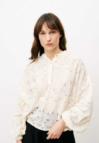 Weiße Bluse aus floraler Spitze mit einem Knopfkragen, langen Ballonärmeln und einem strukturierten Design. Über einem dunklen Kleidungsstück getragen.
