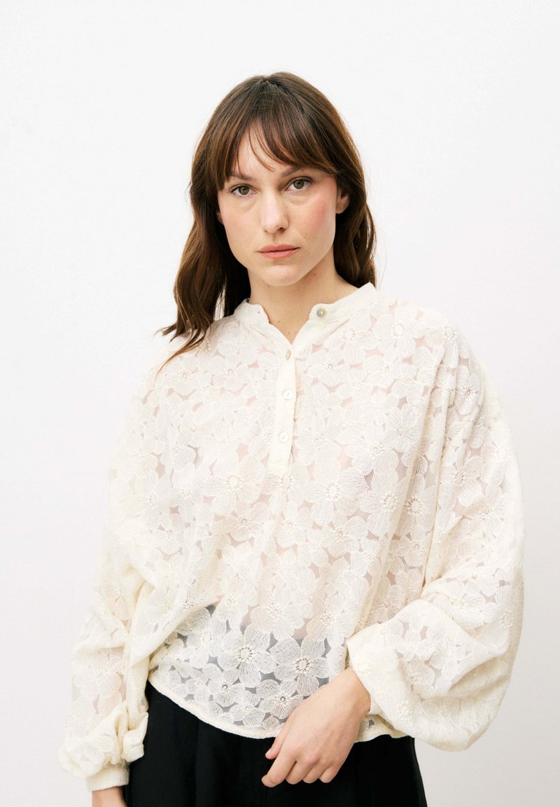 Weiße Bluse aus floraler Spitze mit einem Knopfkragen, langen Ballonärmeln und einem strukturierten Design. Über einem dunklen Kleidungsstück getragen.