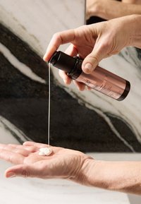 Butelka z pompką z czarną nakrętką, wyciskająca kremowy, biały balsam na dłoń, ustawiona na tle marmuru z szarymi i białymi żyłkami.