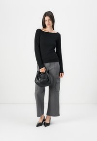 Giovane donna che indossa un top a maniche lunghe nero e pantaloni cargo grigi, tiene in mano una borsa nera ed è in scarpe col tacco nere a punta.