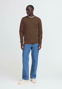 Brauner Strickpullover mit Rundhalsausschnitt, kombiniert mit hellblauen geraden Jeans und weißen Sneakers, vor einem einfarbigen Hintergrund stehend.