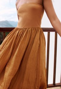 Pántnélküli ruha meleg, visszafogott barna színben. Testhez simuló felsőrész, fodros, légies szoknyával, amely texturált, könnyű anyagból készült.