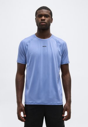 Mlad moški, oblečen v svetlo modro športno majico s kratkimi rokavi z logotipom "BOSS", stoji na belem, enobarvnem ozadju.