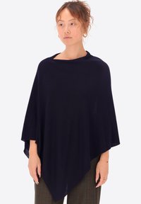 Poncho lavorato a maglia blu navy con un design ampio e drappeggiato, caratterizzato da un colletto rotondo e materiale testurizzato. Indossato sopra pantaloni con fantasia.