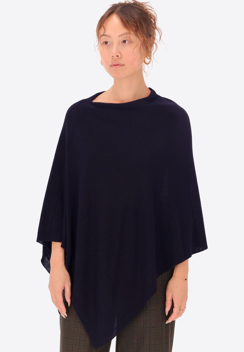 Poncho lavorato a maglia blu navy con un design ampio e drappeggiato, caratterizzato da un colletto rotondo e materiale testurizzato. Indossato sopra pantaloni con fantasia.