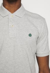 Harmaa polo-paita, jossa on teksturoitu kangas, klassinen kaulus, kaksipainikeinen napitus ja rintataskussa vihreä brodeerattu puu-logo.