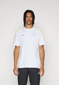 Witte kortslippige jersey met gele schouderstrepen, groene logo-accenten en rode stiksels. Gecombineerd met zwarte broeken met een groen logo.
