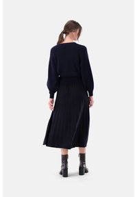 Marineblauer Strickpullover mit Puffärmeln, kombiniert mit einem plissierten marineblauen Rock und Leder-Stiefeletten mit quadratischem Absatz, von hinten betrachtet.