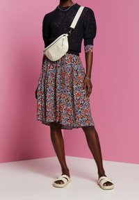 Nainen, joka pukeutuu mustaan neulepaitaan, kukkakuosisesti hameeseen, valkoiseen olkalaukkuun ja valkoisiin sandaaleihin, seisoo vaaleanpunaisen taustan edessä.