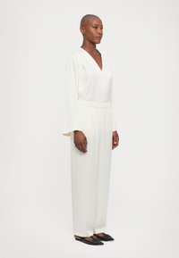 Mono de marfil con mangas largas, escote en V, cinturilla elástica y diseño de pierna ancha, hecho de material suave y ligero. Combinado con zapatos planos negros.