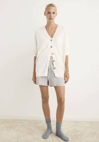 Cardigan bianco con bottoni, abbinato a pantaloni corti grigi a righe. La modella indossa calze grigie. Tessuto morbido, vestibilità comoda, design a V.