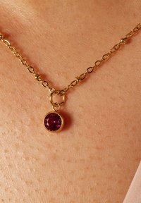 Goldene Halskette mit einem runden, facettierten lila Steinanhänger mit polierter Oberfläche, der auf strukturiertem Haut ruht.