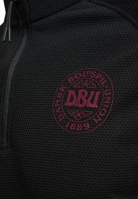 Svart texturerad tyg med maroon cirkulärt logotyp som står "DANSK BOLDSPIL-UNION 1889" och initialerna "DBU" i centrum.