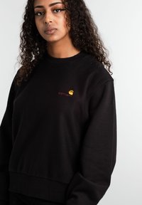 Zwarte sweatshirt van katoen met een ribgebreide zoom en manchetten. Heeft een klein oranje geborduurd logo op de borst. Eenvoudig, klassiek ontwerp.