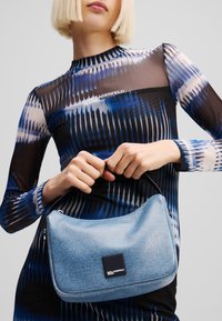 Denim handbag in light blue with a smooth texture, semi-structured shape, and a small black logo patch. Model wears a patterned long-sleeve top.