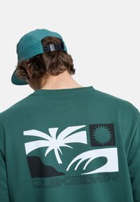 Sudadera verde oscuro con un diseño gráfico blanco de palmeras, sol y olas. La parte trasera tiene un logo en una tipografía elegante. Gorra azul aguamarina en el fondo.
