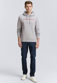 Pilkos spalvos gobtuvu megztinis su raudonu "TOM TAILOR" logotipu, derinamas su tamsiai mėlynais džinsais ir baltais sportbačiais. Lygios medžiagos, laisvo kirpimo dizainas.