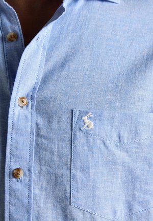 Hellblaues Hemd mit Knopfleiste und Brusttasche, die ein kleines weißes, gesticktes Kaninchenlogo zeigt.