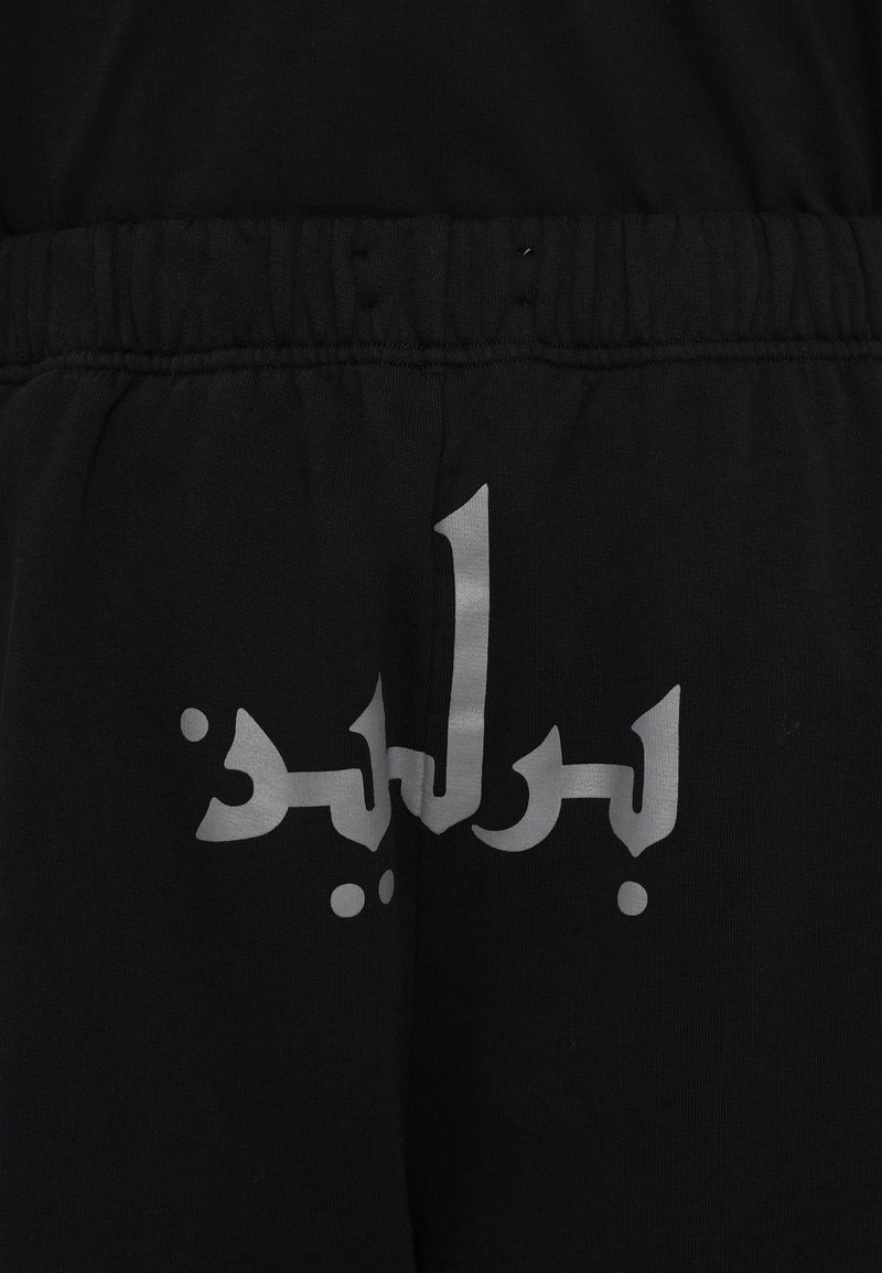 Joggers negros con texto árabe plateado en la parte posterior, hechos de suave material de algodón con una cintura elástica para mayor comodidad.