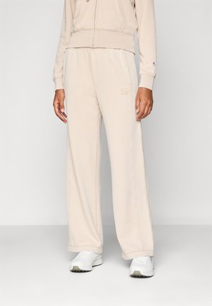 Person trägt beige weite Jogginghose und passenden Reißverschluss-Hoodie, kombiniert mit weißen Sneakers, steht vor einem einfarbigen Hintergrund.
