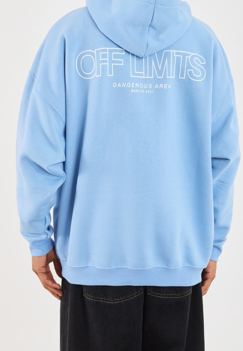Jasnoniebieska bluza z kapturem z dużym białym napisem na plecach: "OFF LIMITS DANGEROUS AREA BERLIN 2001". Miękki materiał.