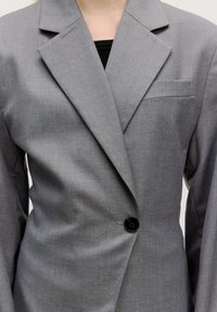 Grijze op maat gemaakte blazer met een gekarteld kraag, enkele zwarte knoopsluiting en een borstzak. De stof heeft een gladde, gestructureerde afwerking.