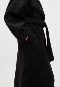 Cappotto in pile nero con superficie texturizzata, maniche lunghe e cintura coordinata. Presenta tasche laterali e colletto con bottoni.