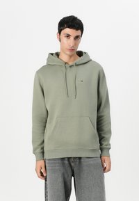 Green hoodie made of soft fabric, featuring a front pocket, adjustable drawstring hood, and a small logo on the chest. Worn with gray jeans.