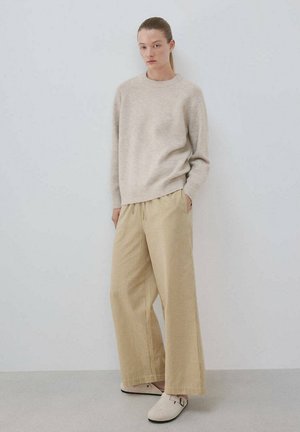 Mujer con coleta que lleva un suéter beige, pantalones anchos beige y zapatos claros sin cordones, de pie contra una pared clara y lisa.