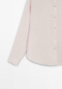 Světle růžová lněná košile s dlouhým rukávem, manžetami na knoflíky a knoflíky vepředu, zobrazená od poloviny těla po levý rukáv.