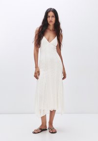Abito bianco a lunghezza ginocchio con corsetto aderente, scollatura a V e motivo testurizzato. La modella indossa infradito nere, in piedi davanti a uno sfondo neutro.