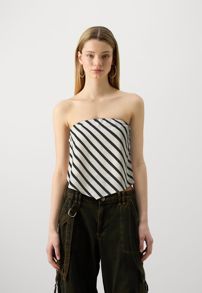 Haut sans bretelles en tissu blanc avec des rayures diagonales bleu marine, présentant un ourlet arrondi. Associé à un pantalon cargo vert foncé et des boucles d'oreilles créoles.