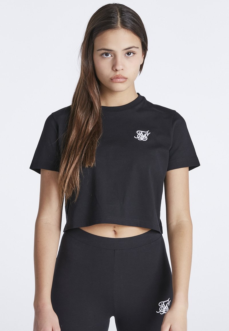 Mladá žena s dlouhými hnědými vlasy, oblékající černé zkrácené tričko a odpovídající černé legíny s bílými vyšívanými logy.