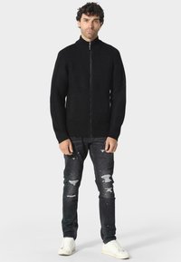 Černá zipová mikina s ribrovou strukturou, vysokým límcem a dvěma bočními kapsami, spárována s roztrhanými tmavými džíny a bílými teniskami.