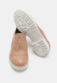 Chaussures en cuir tan à enfiler avec des détails brogue et de épaisses semelles en caoutchouc blanc, une chaussure montrant un motif de semelle détaillé en dessous.