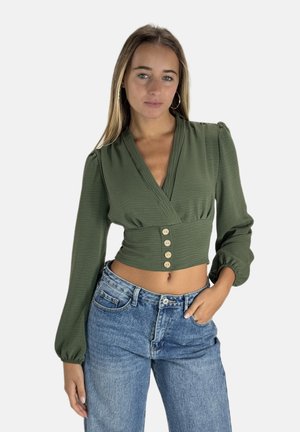 Blusa corta verde oliva con design a portafoglio, maniche puff, e cinque bottoni di legno sul davanti. Abbinata a jeans blu a vita alta.